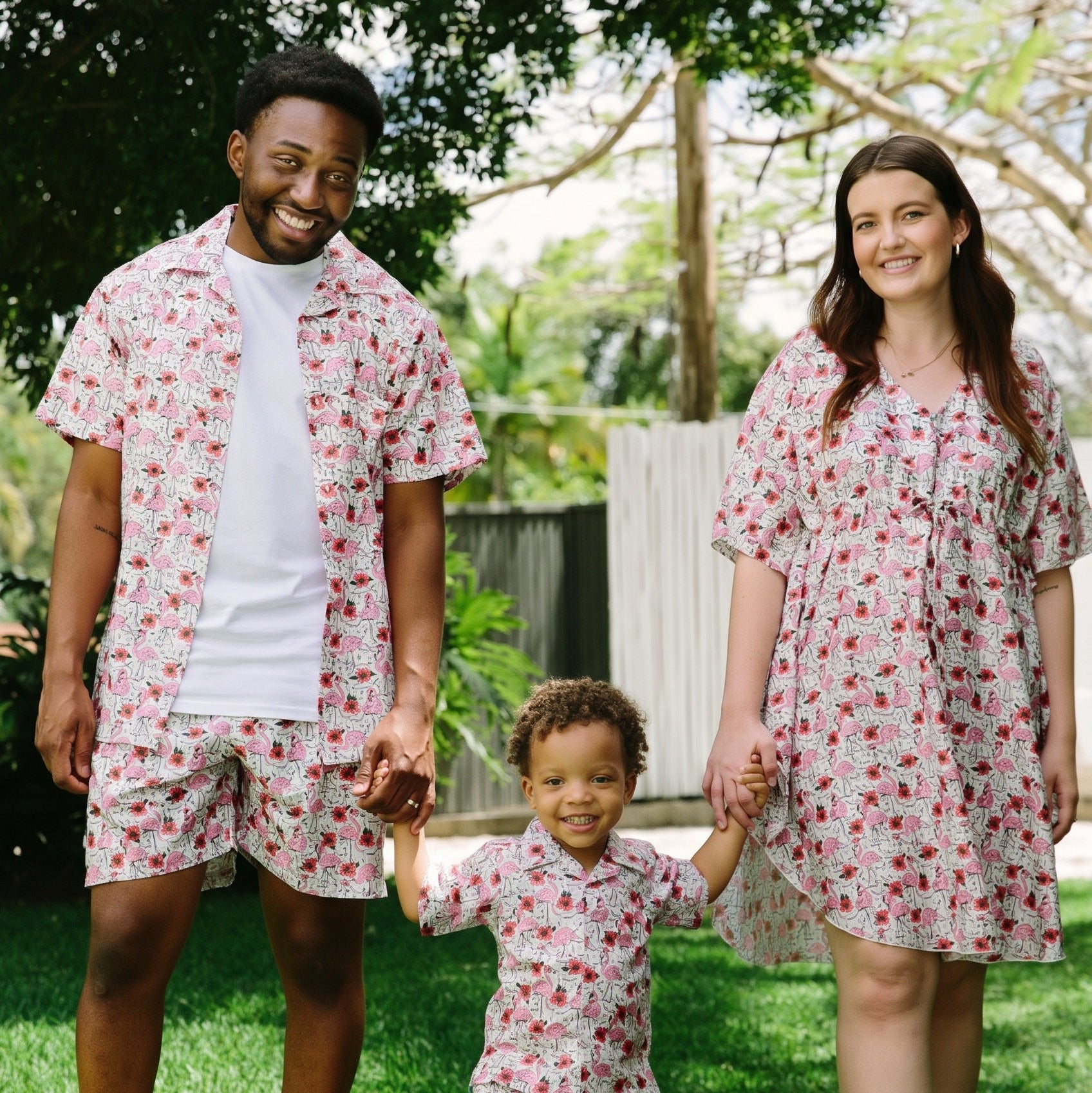 A family of three wearing matching floral-print loungewear sets stands on a grassy lawn outdoors. The set includes a short-sleeve button-up shirt and shorts for the adults, and a matching tunic dress and shorts set for the child.