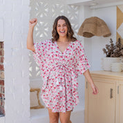 A woman modeling a short, loose white kaftan dress with a red and pink floral print, standing in a bright, modern room with white brick walls and wooden floors.