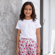A young girl stands outdoors wearing a white t-shirt and pink floral print shorts with a white drawstring, posing in front of a gray marble wall and a white sliding door.