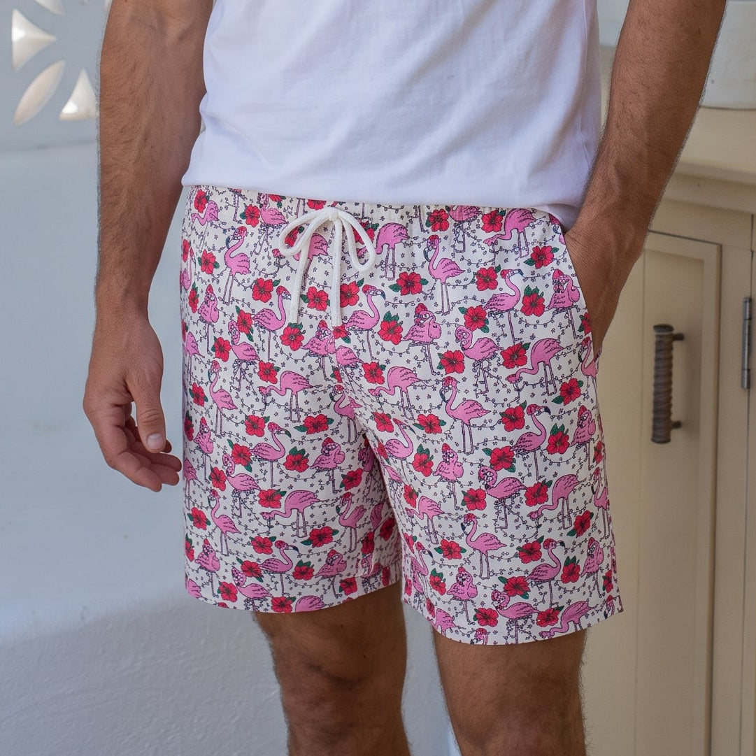 A man wearing a white crewneck t-shirt and pink and white patterned swim shorts, standing in a bright, modern bathroom setting with holiday decorations.