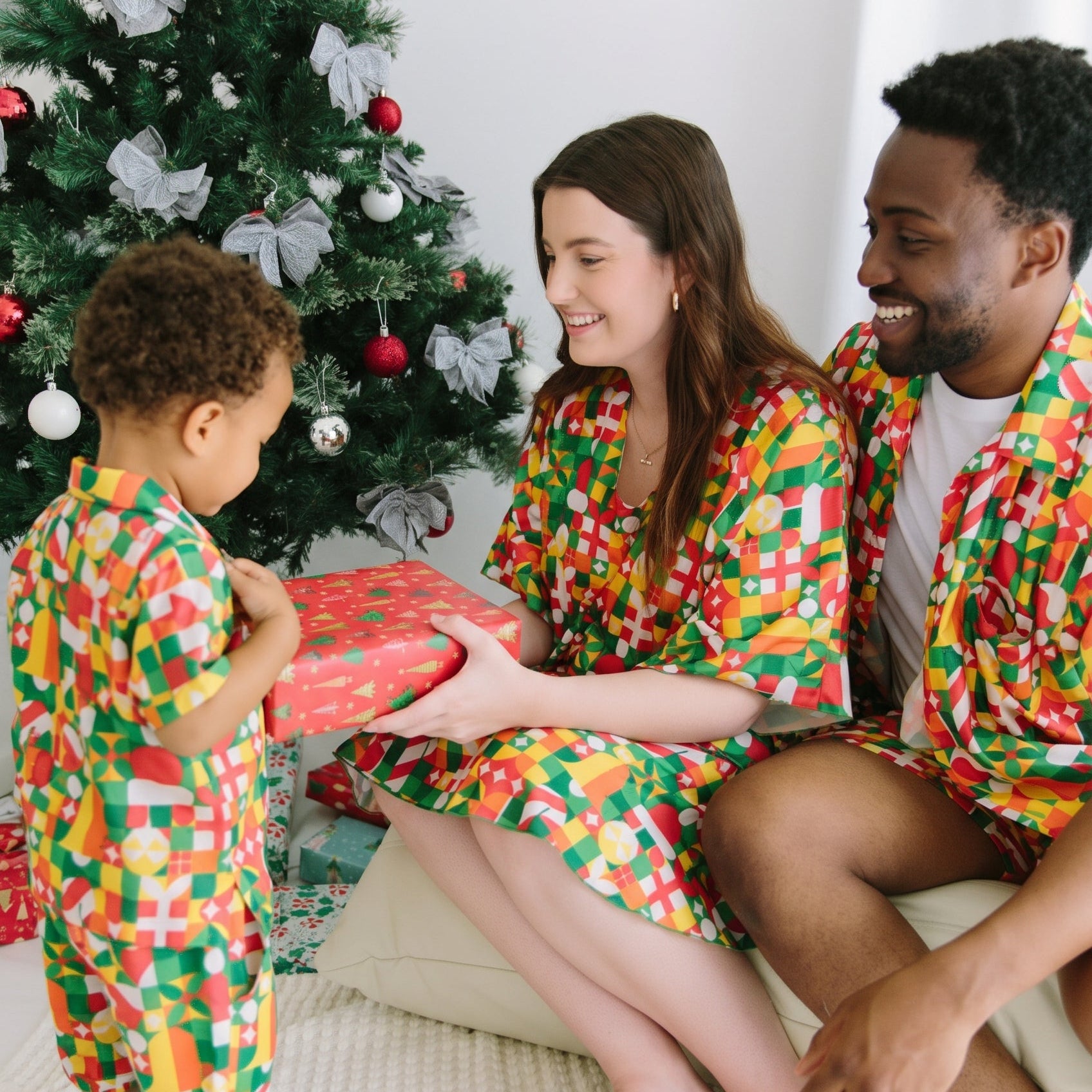 A family of three wearing matching colorful geometric print pajamas and bathrobes sits on a white sofa in front of a decorated Christmas tree, with the woman holding a wrapped gift.