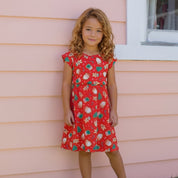 A young girl with curly light brown hair stands outdoors against a peach-pink house wall, wearing a red knee-length dress with a festive holiday print of green pine trees, white ornaments, and gold stars. She is wearing white sneakers.
