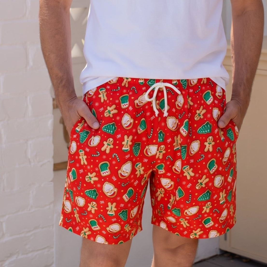 A man wearing red patterned swim trunks with a white drawstring, a plain white t-shirt, and black sunglasses, standing in a bright, modern indoor setting.