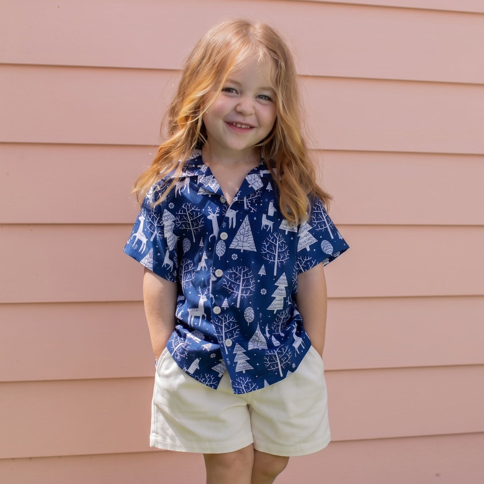 A young girl with long red hair stands barefoot on a green lawn in front of a pink house siding, wearing a blue short-sleeve button-up shirt with a white tree and snow pattern, paired with cream shorts.