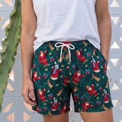 A woman stands outdoors in front of a white decorative fence and cactus, wearing dark green holiday print shorts with a white drawstring, a white sleeveless top, and beige slide sandals.