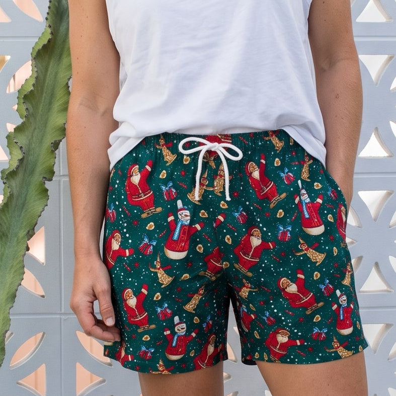 A woman stands outdoors in front of a white decorative fence and cactus, wearing dark green holiday print shorts with a white drawstring, a white sleeveless top, and beige slide sandals.
