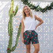 A woman stands outdoors in front of a white decorative lattice wall and a tall cactus, wearing a white sleeveless top and dark green shorts with a red and yellow tropical floral print and white drawstring.
