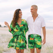 A couple walking on a sandy beach wearing matching tropical print swimwear. The woman wears a green palm-patterned short dress, and the man wears matching green swim shorts with a white drawstring and a white short-sleeve button-up shirt.