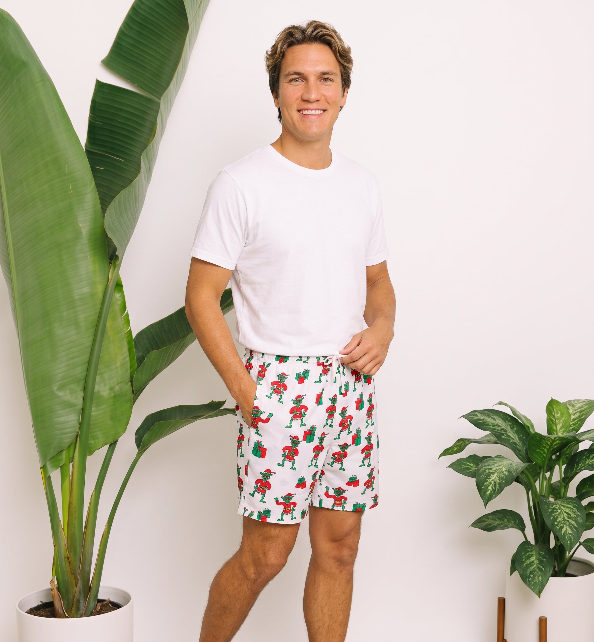 A man wearing a white t-shirt and white shorts with a red and green Santa Claus pattern, standing in a brightly lit room with potted plants.