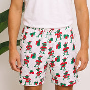 A man wearing a white crewneck t-shirt and white drawstring shorts with a green and red Santa Claus pattern, standing in front of a white wall with tropical plants.