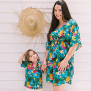 A woman and a child wearing matching tropical floral Hawaiian-style outfits, standing barefoot against a white wood-paneled wall with a large straw hat hanging above them.