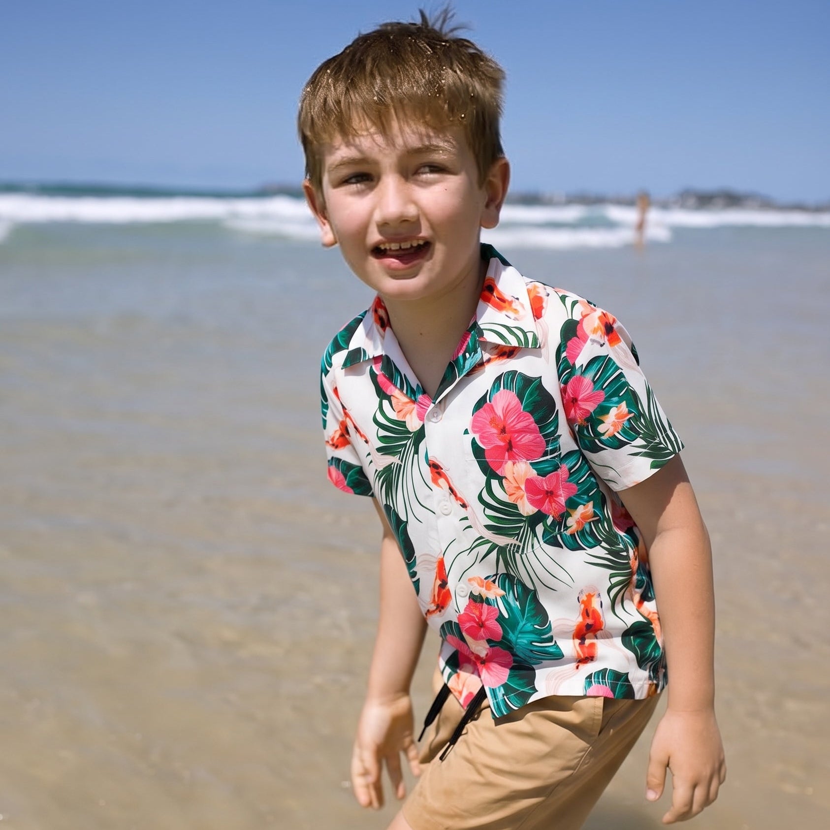 A young boy smiling on a sunny beach, wearing a white short-sleeve Hawaiian-style button-up shirt with a vibrant green, pink, and orange tropical floral print, paired with tan pants.