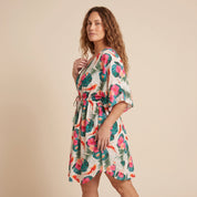A woman modeling a short tropical print dress with pink hibiscus flowers and green palm leaves on a cream background, set against a solid beige studio backdrop.