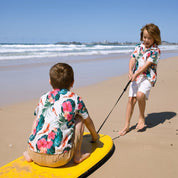 Two children in matching tropical floral shirts and white shorts on a sandy beach, with one sitting on a yellow board and the other standing and holding a leash.