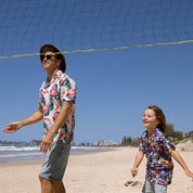 A man and a young boy walking and smiling on a sunny beach, both wearing colorful floral short-sleeve button-up shirts. A volleyball net is visible in the foreground, with the ocean and sky in the background.