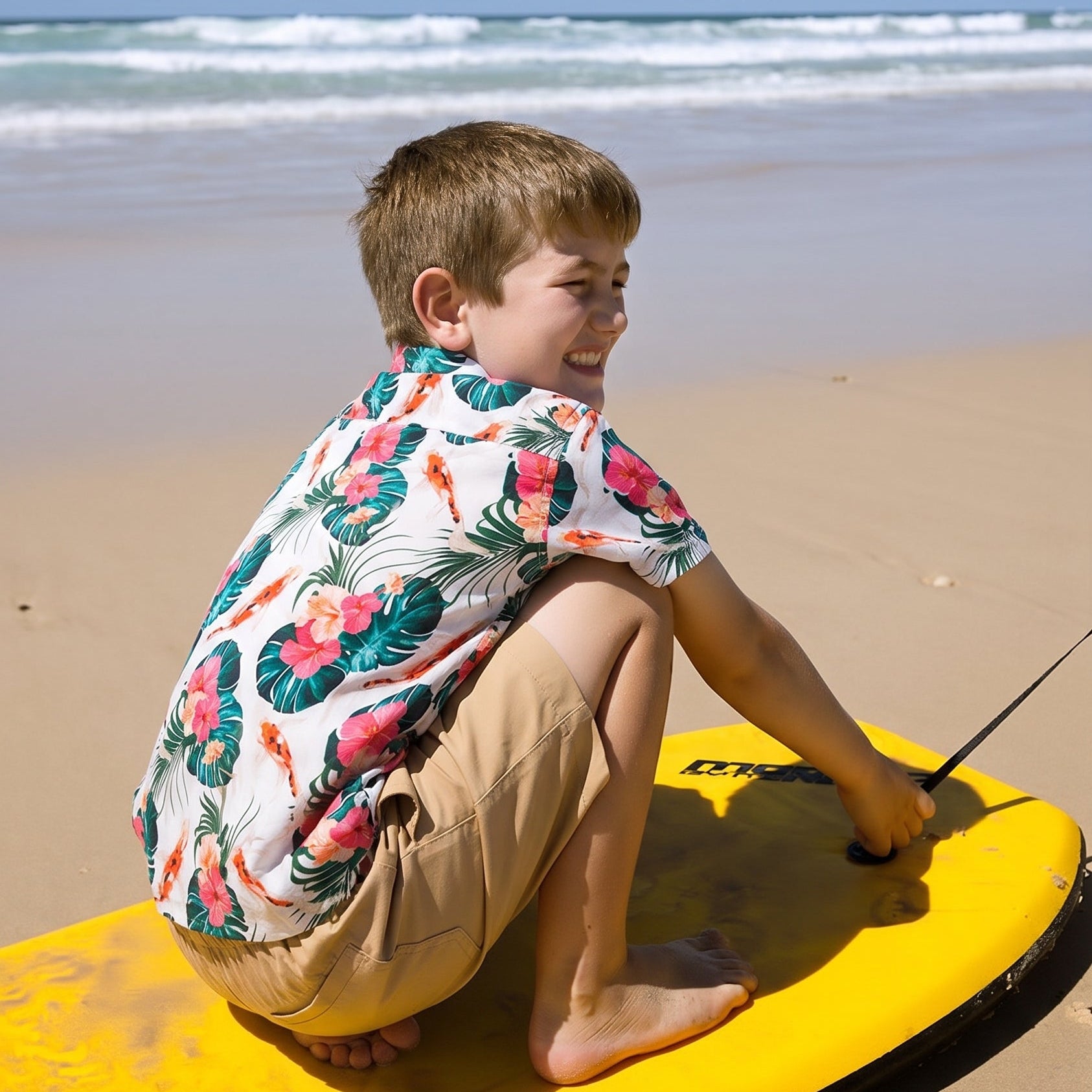 A young boy wearing a colorful tropical print short-sleeve shirt and khaki shorts sits on a yellow bodyboard on a sandy beach with the ocean and blue sky in the background.