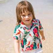 A young child standing in shallow water on a beach, wearing a white short-sleeve button-up shirt with a colorful tropical print featuring green leaves, pink hibiscus flowers, and orange fish.