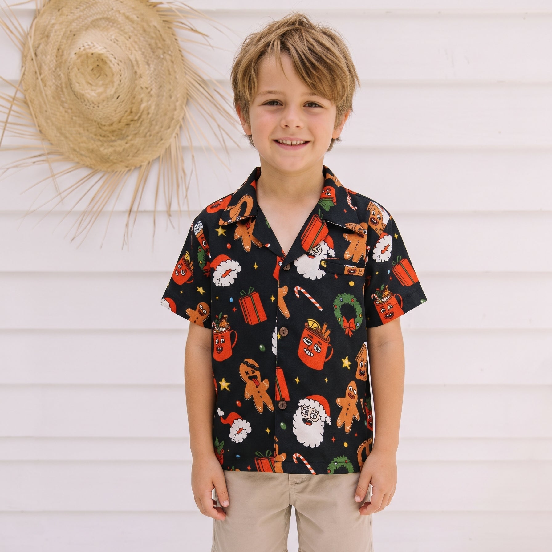 A young boy stands against a white wooden wall wearing a black short-sleeve button-up shirt with a colorful Christmas-themed print, paired with khaki shorts.