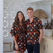 A smiling couple stands indoors wearing matching dark short-sleeve holiday-themed shirts and a matching robe. The fabric has a festive print with Santa Claus, pumpkins, ornaments, and green wreaths.