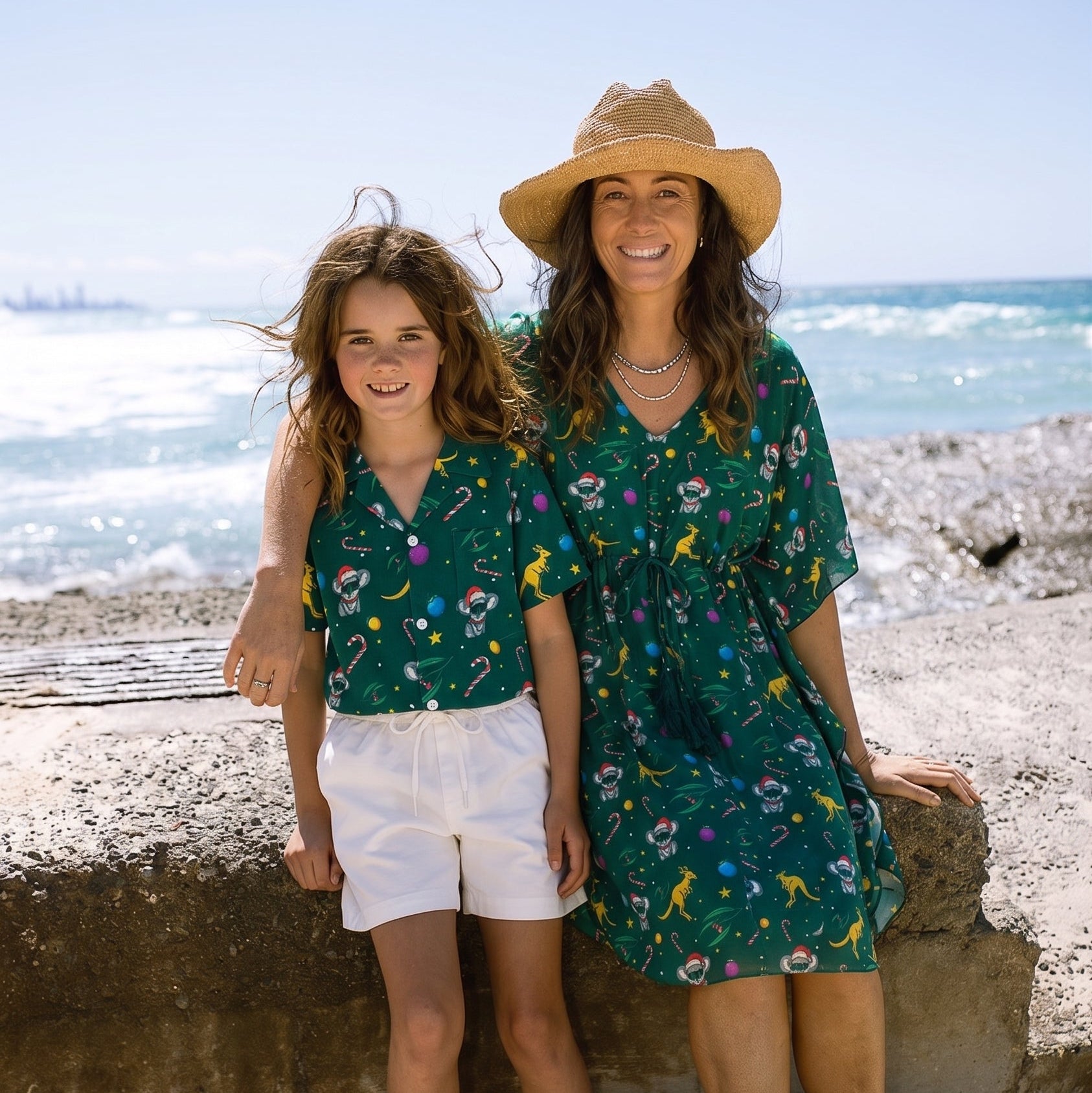 A woman and a girl standing together on a sandy beach in front of the ocean. The woman wears a wide-brim straw hat and a green floral dress, and the girl wears a matching green floral top and white shorts.