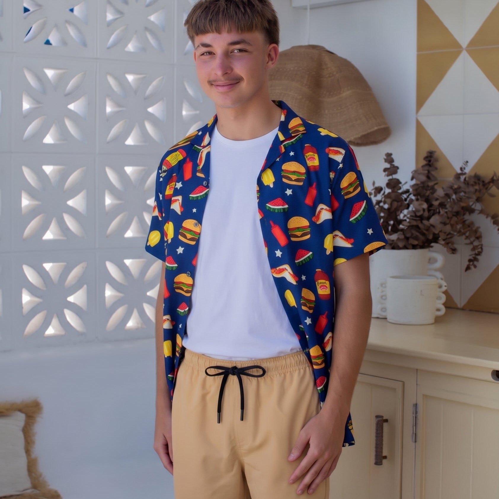 A young man wearing a blue short-sleeve button-up shirt with a colorful burger print, a white t-shirt, and beige shorts, standing in a bright, modern room with white walls and decorative panels.
