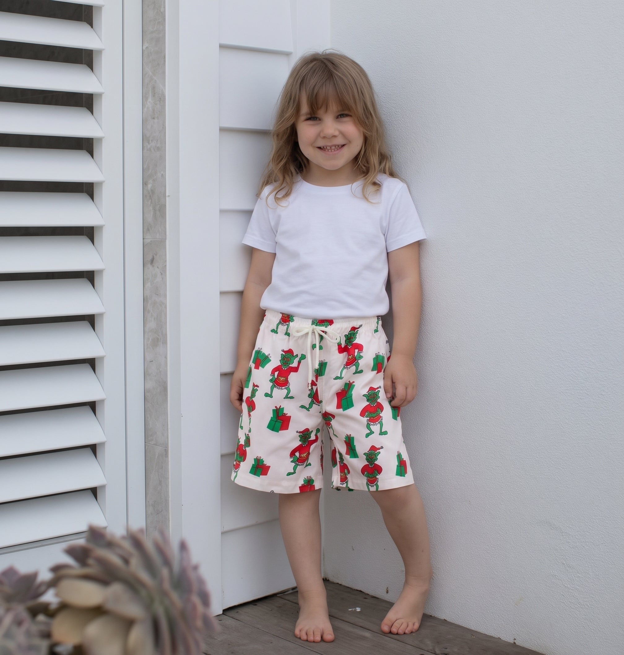 A young girl with light brown hair stands barefoot against a white wall, wearing a white t-shirt and cream-colored shorts with a red and green Santa Claus pattern.