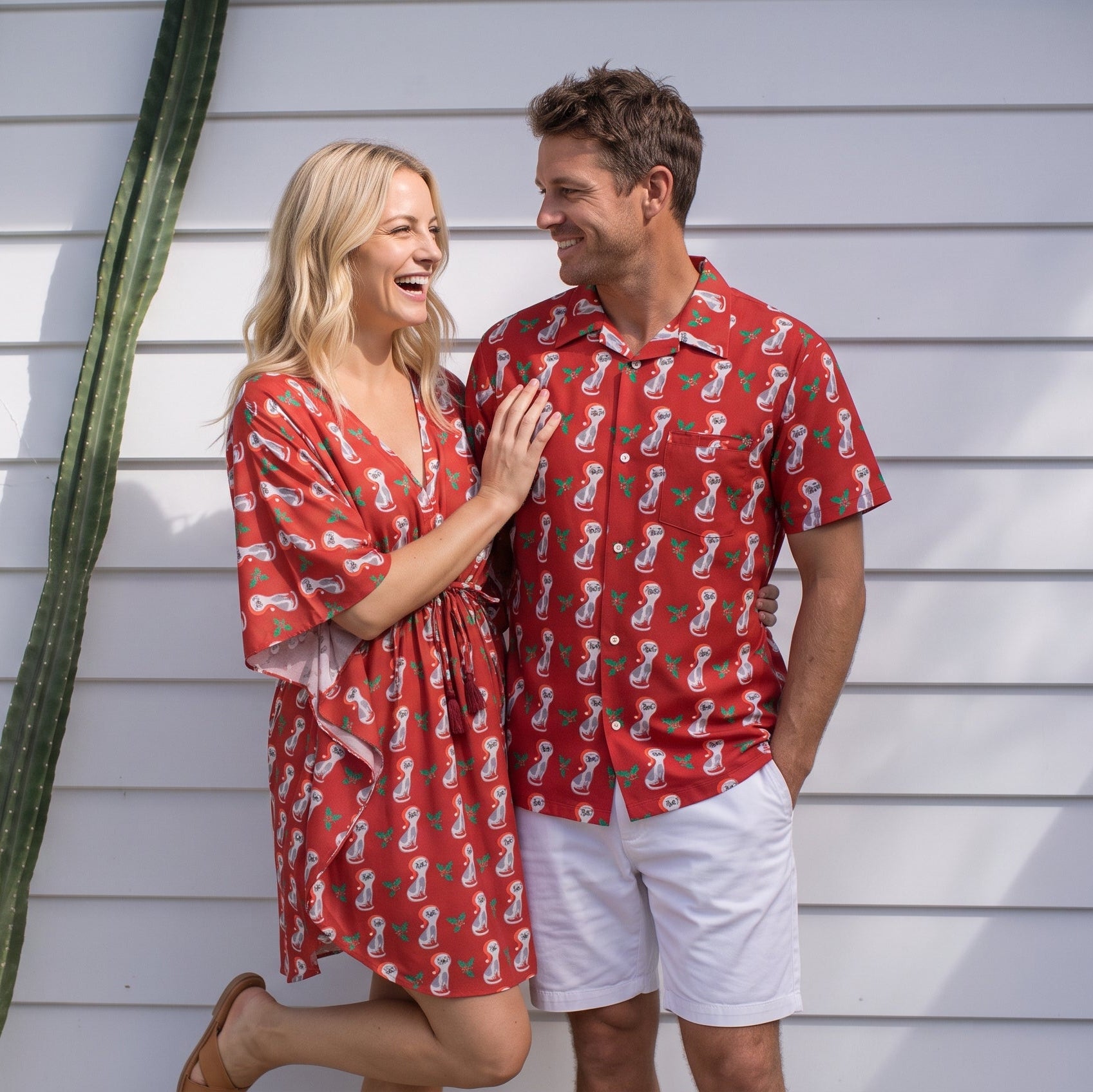 A man and a woman wearing matching red and white patterned resort wear, standing in front of a white house wall with a cactus on the left.
