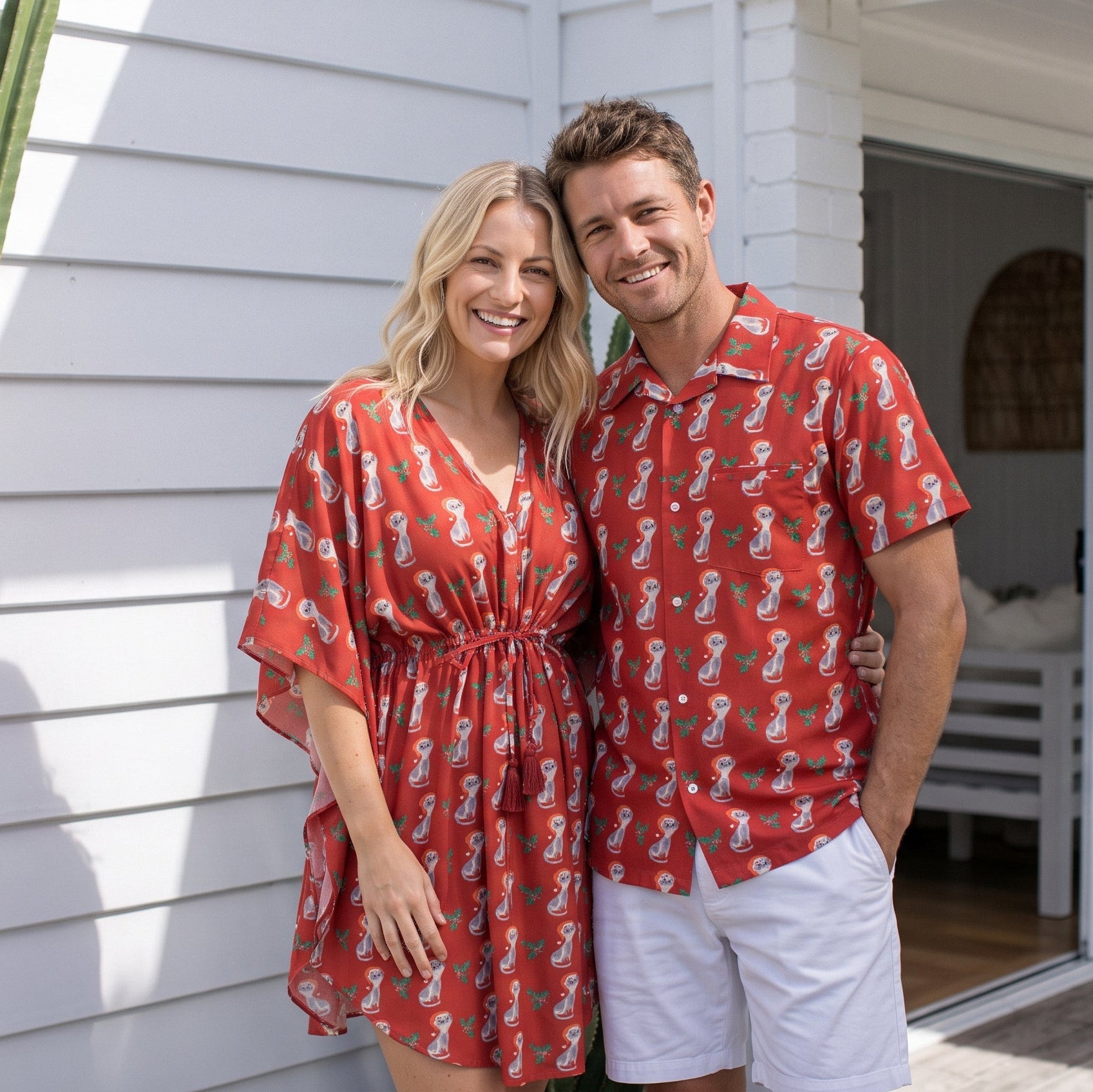A smiling couple standing outdoors in front of a white house. The woman wears a red short kaftan dress with a white paisley print, and the man wears a matching red short button-up shirt with the same print, paired with white shorts.