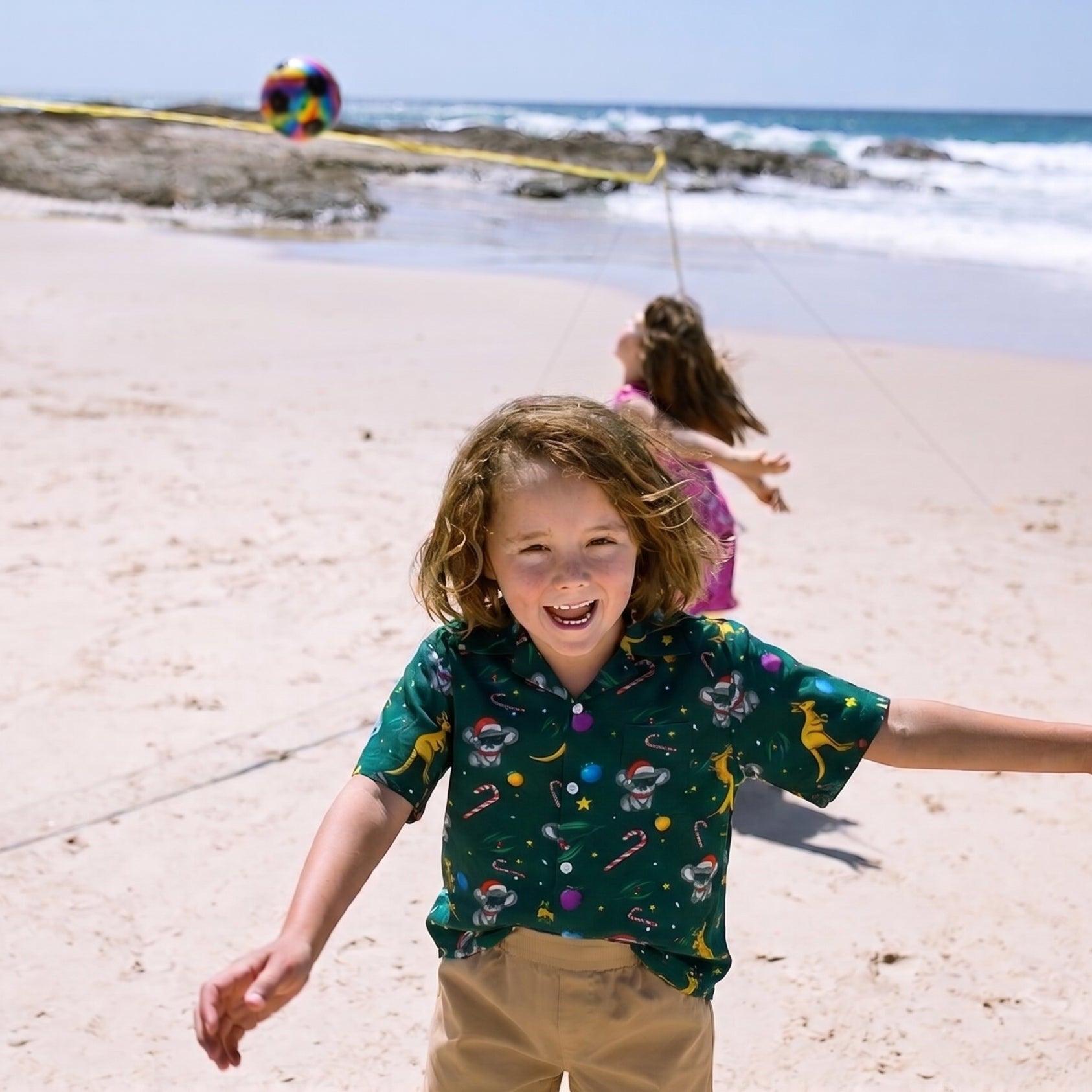 A young boy with curly brown hair runs and smiles on a sandy beach, wearing a dark green short-sleeve button-up shirt with a colorful candy cane and space-themed print, paired with khaki pants. Other children and a rocky shoreline are visible in the background.