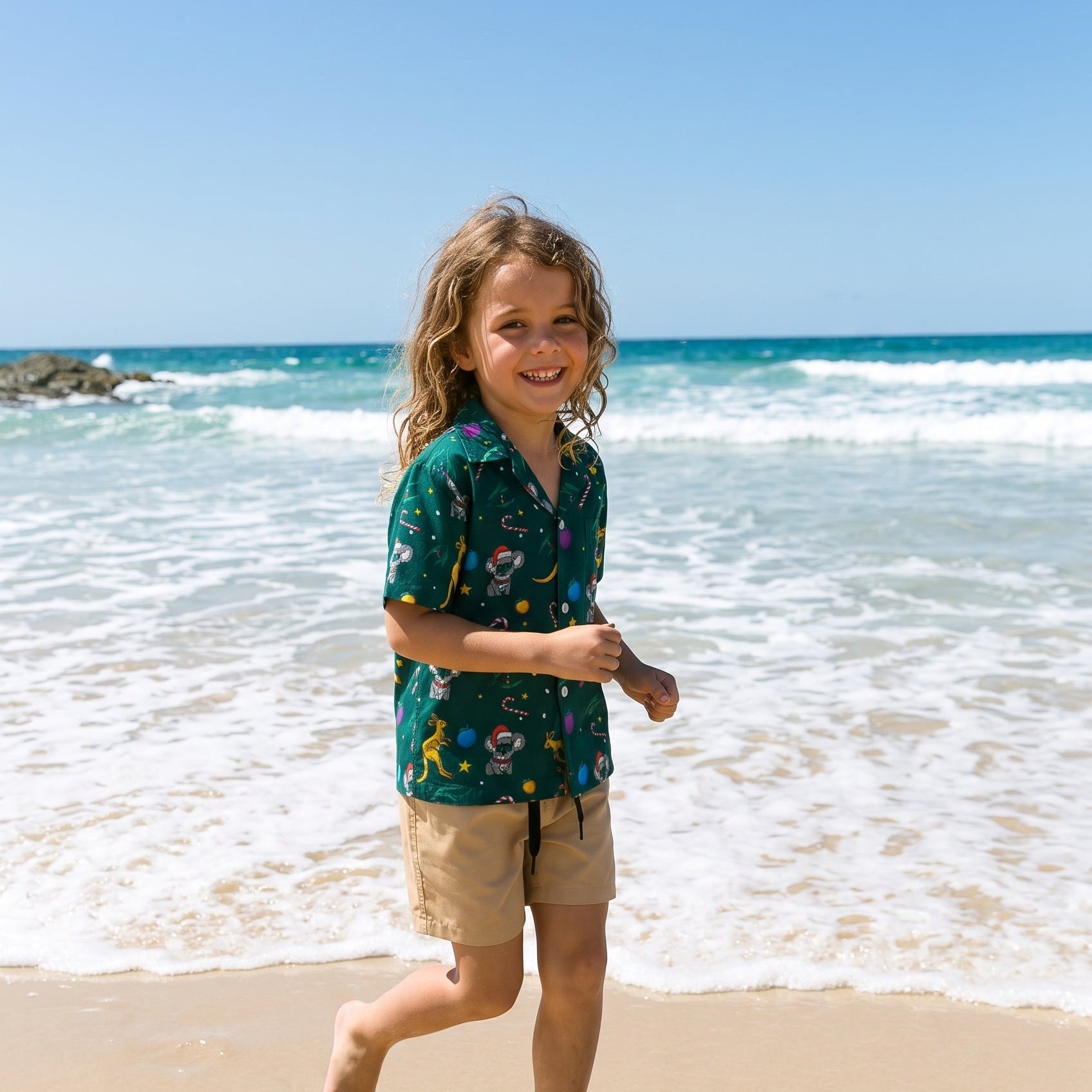 A young boy running on a sandy beach near the ocean waves, wearing a dark green short-sleeve button-up shirt with a colorful tropical floral print and tan shorts.
