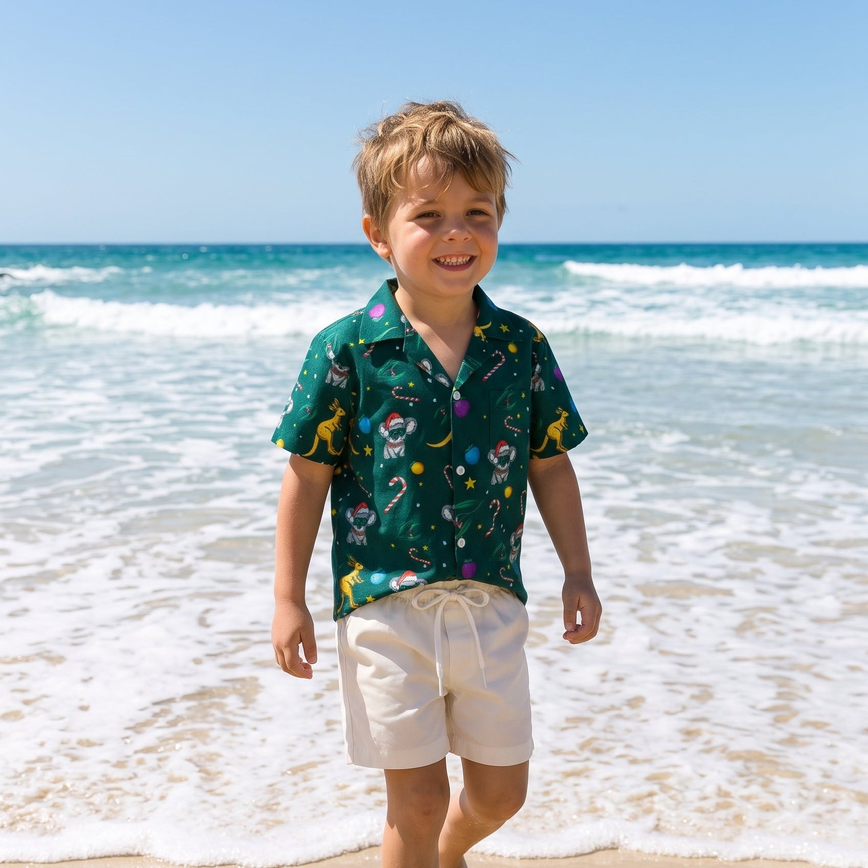 A young boy walking barefoot on a sandy beach with ocean waves behind him, wearing a dark green short-sleeve button-up shirt with a colorful space-themed print and off-white shorts.