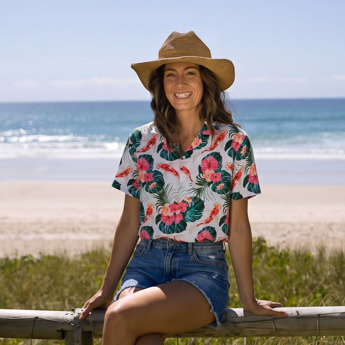 A woman wearing a straw hat, a white short-sleeve Hawaiian shirt with a red and green floral print, and denim shorts sits on a wooden fence overlooking a beach.