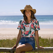 A woman wearing a straw hat, a white short-sleeve Hawaiian shirt with a red and green floral print, and denim shorts sits on a wooden fence overlooking a beach.