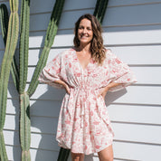 A woman wearing a light pink and white floral print mini dress with wide sleeves, standing outdoors against a white siding wall next to a tall cactus.