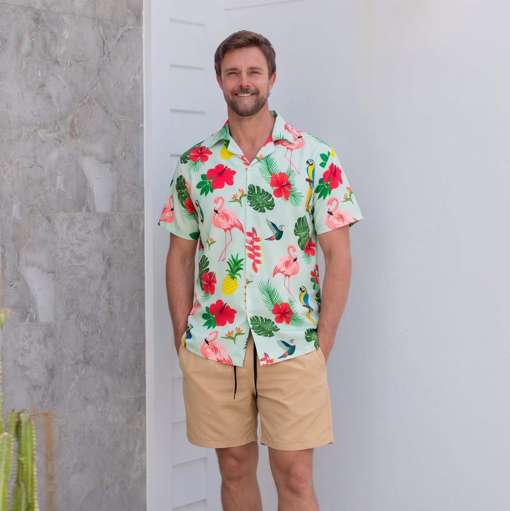 A man wearing a short-sleeve Hawaiian-style camp shirt with a tropical floral and flamingo print, paired with khaki shorts and black slide sandals, standing in a modern indoor setting with a white wall and textured gray stone column.
