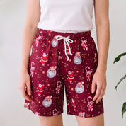 A woman stands indoors wearing a white sleeveless tank top and burgundy holiday print shorts with a white drawstring. Large green potted plants are visible on both sides against a white wall.