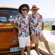 Two men wearing matching tropical floral short-sleeve button-up shirts, tan shorts, and straw hats posing in front of an orange vintage Volkswagen bus outdoors.