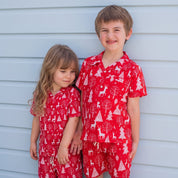 Two children wearing matching red holiday pajama sets with a white Christmas tree and reindeer print, standing barefoot against a light blue siding wall.