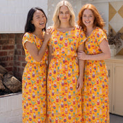 Three women standing together in matching yellow maxi dresses with a colorful cartoon character print, set against a bright, modern outdoor patio background.