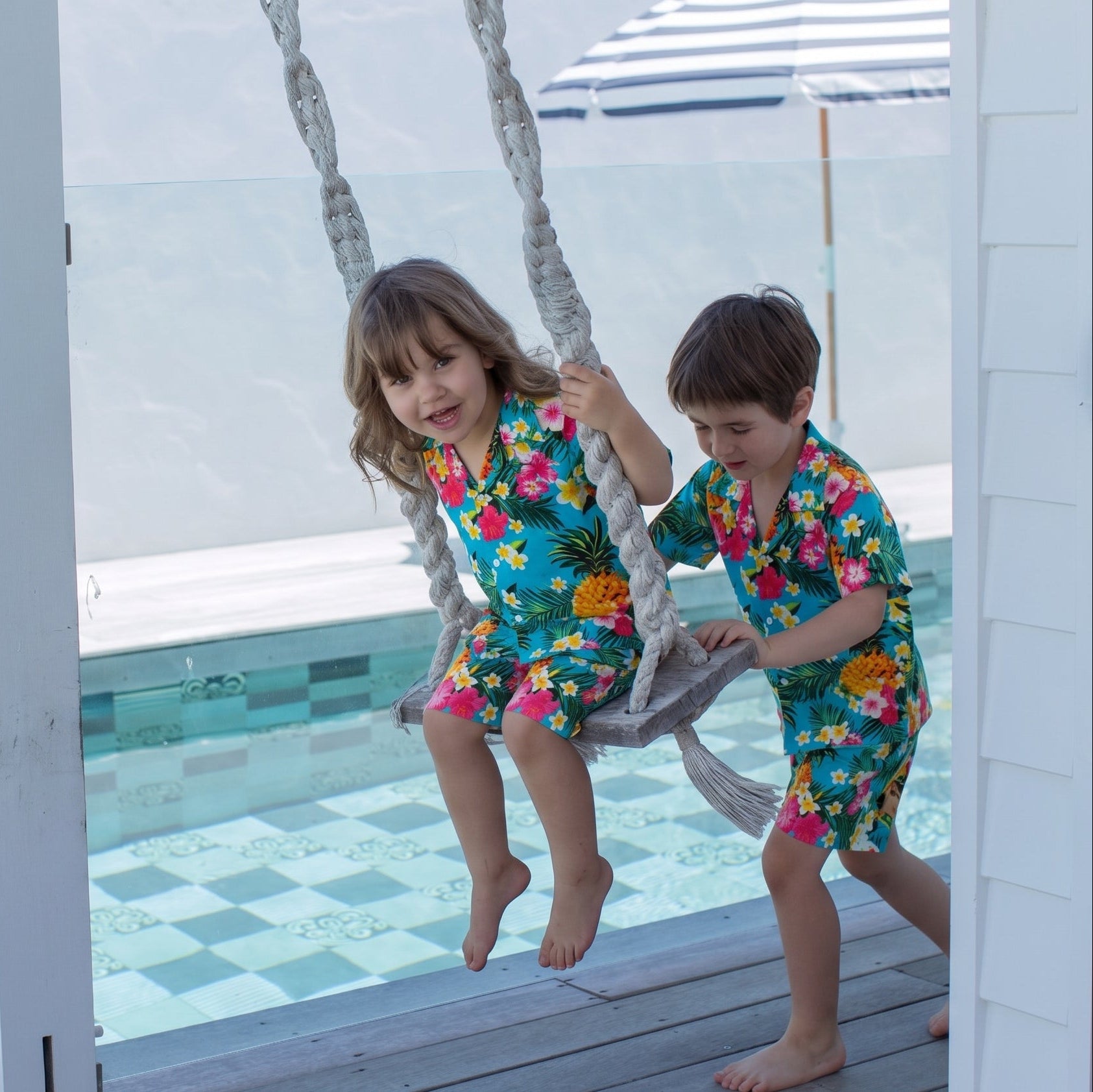 Two children wearing matching floral swim shirts and shorts play on a rope swing by a swimming pool.