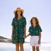 A woman and a young girl standing hand-in-hand on a sunny beach. The woman wears a straw hat and a green floral dress, and the girl wears a matching green floral short-sleeve top and white shorts.
