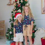 Two young girls standing barefoot in front of a decorated Christmas tree, wearing matching navy short-sleeve shirts with a colorful holiday print and tan shorts. One girl wears a red Santa hat, and wrapped gifts and a white sack sit on the floor around the tree.