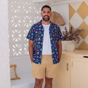 A man stands barefoot in a bright, modern room wearing a short-sleeve button-up shirt with a colorful tropical print, a white t-shirt, and khaki shorts.