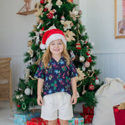 A young child wearing a navy blue Christmas-themed short-sleeve button-up shirt with a colorful holiday print, white shorts, and a red Santa hat, standing barefoot in front of a decorated Christmas tree with wrapped gifts.
