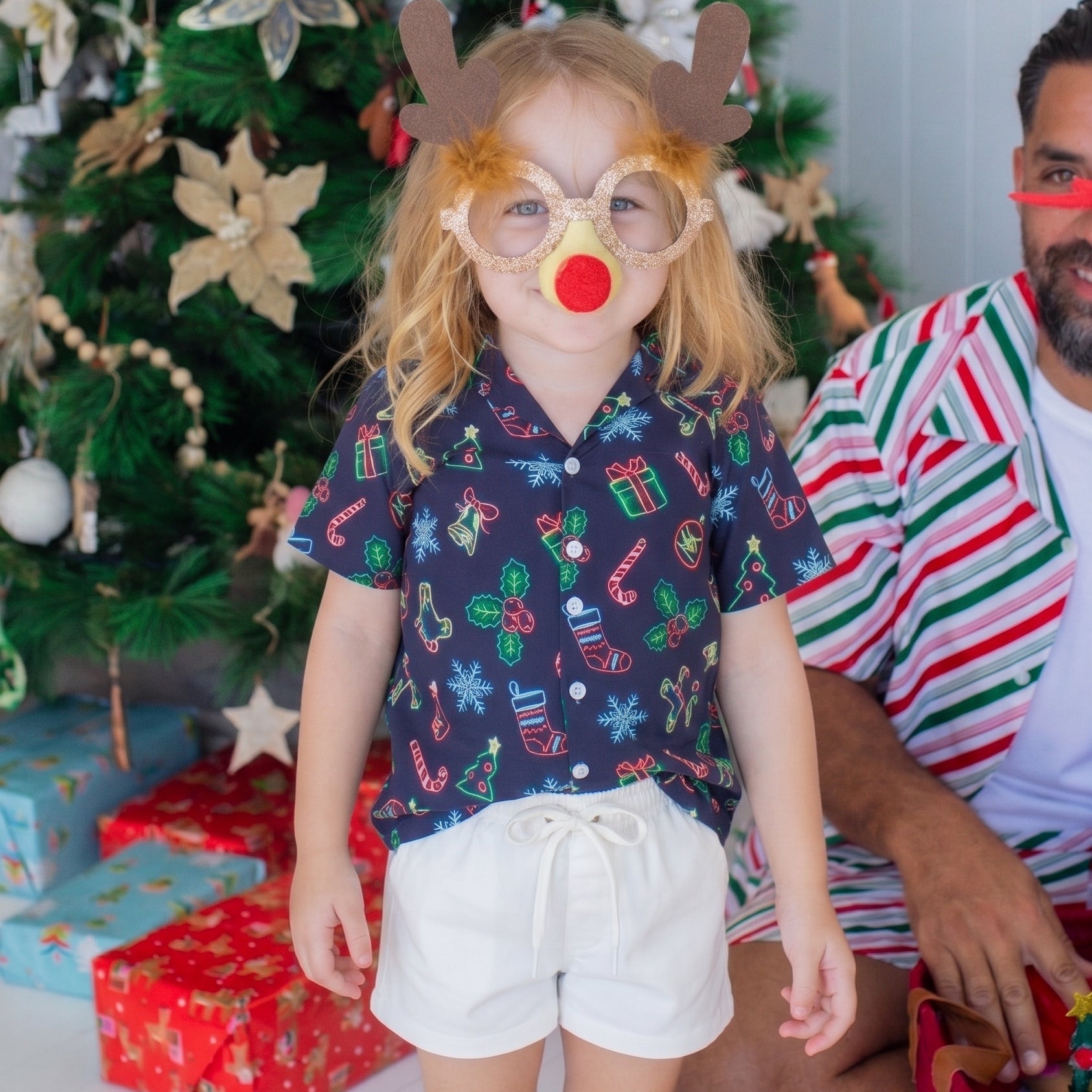 A young girl wearing a Christmas-themed novelty eyeglasses and antler headband with a red nose stands in front of a decorated Christmas tree, with a man in a striped shirt sitting beside her.
