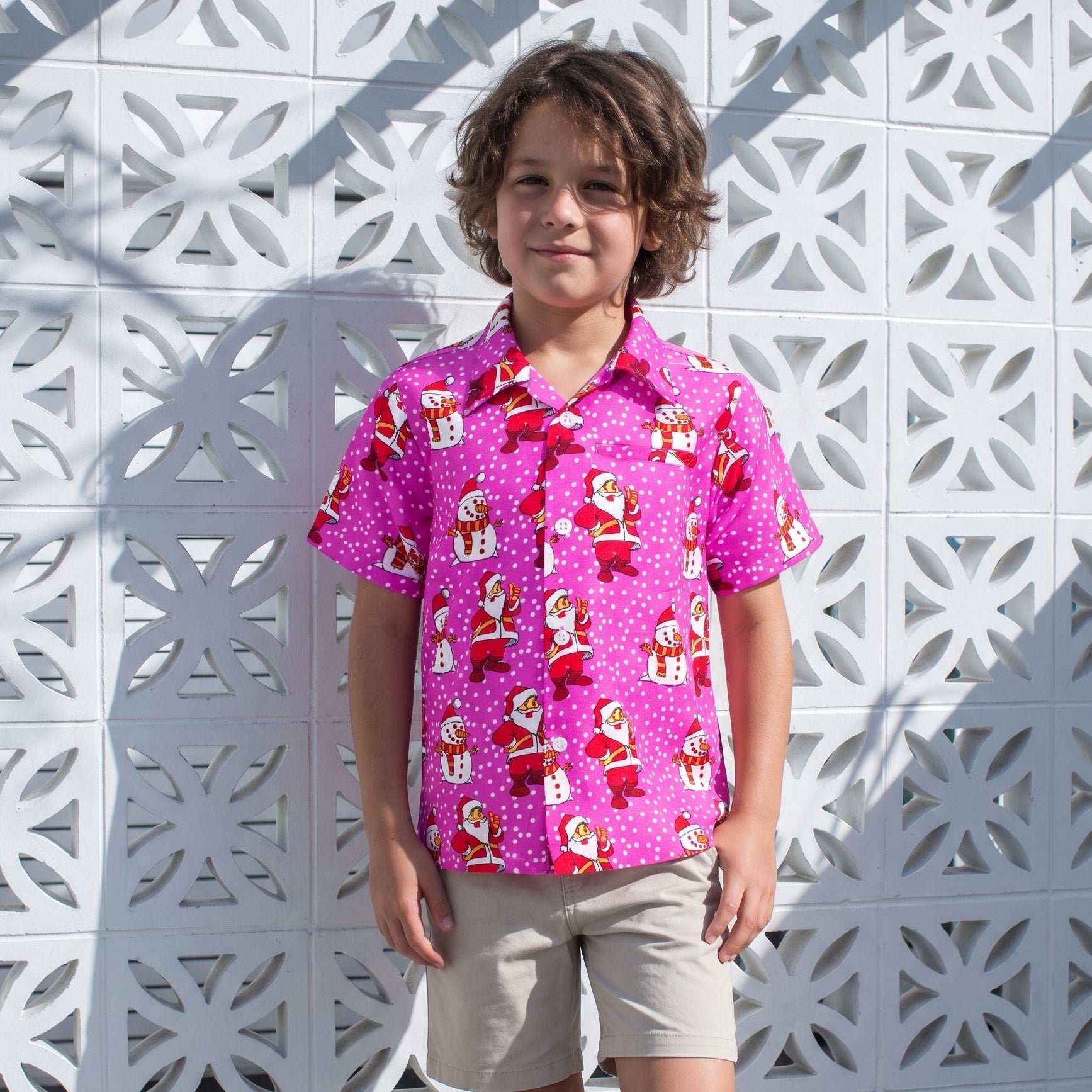 A young boy stands in front of a white geometric patterned wall, wearing a bright pink short-sleeve button-up shirt with a red and white Santa Claus print, paired with khaki shorts and white sneakers.