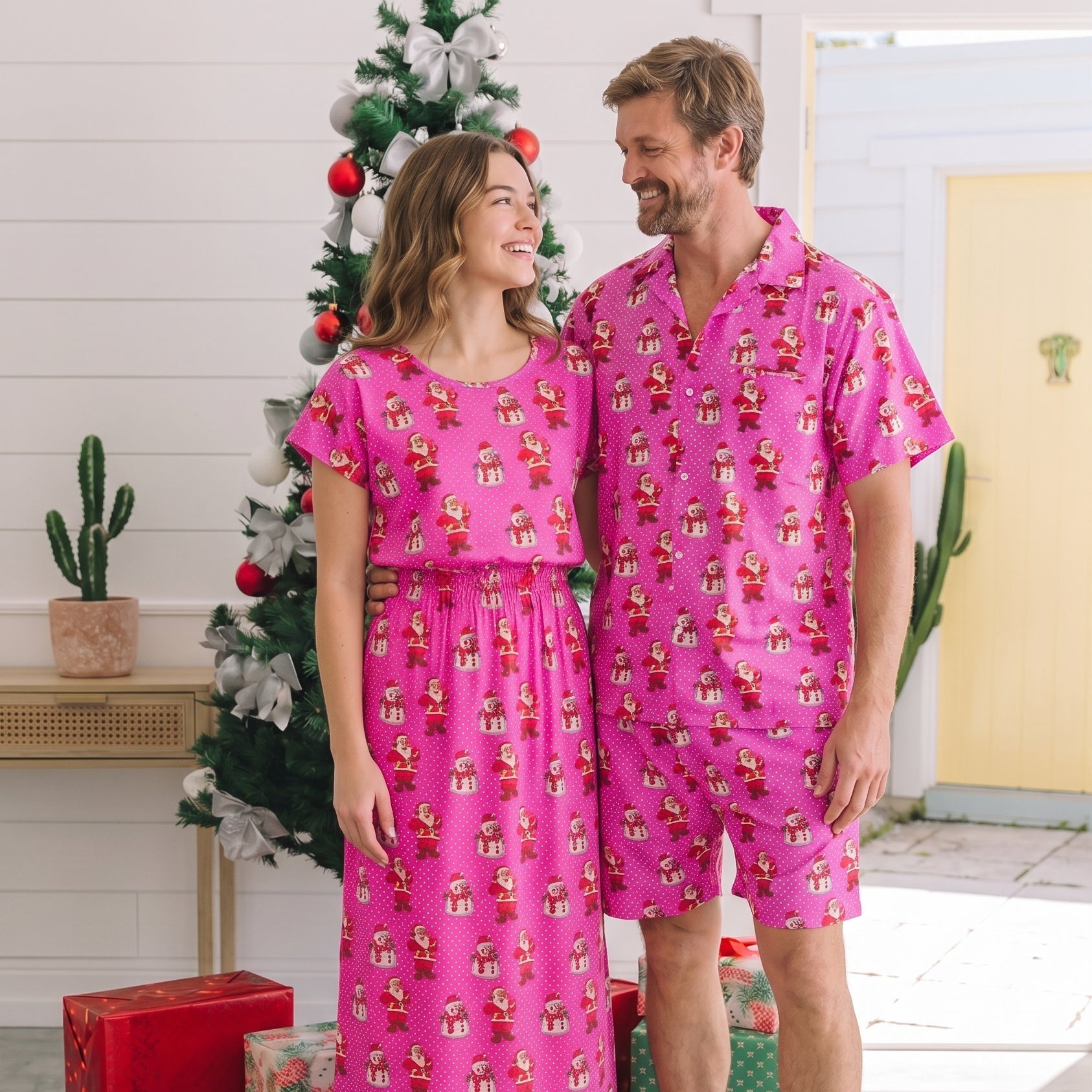 A man and a woman standing together in a bright indoor setting, wearing matching bright pink pajama sets with a repeating Christmas gift print. A decorated Christmas tree, wrapped presents, and indoor decor are visible in the background.