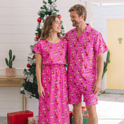 A man and a woman standing together in a bright indoor setting, wearing matching bright pink pajama sets with a repeating Christmas gift print. A decorated Christmas tree, wrapped presents, and indoor decor are visible in the background.