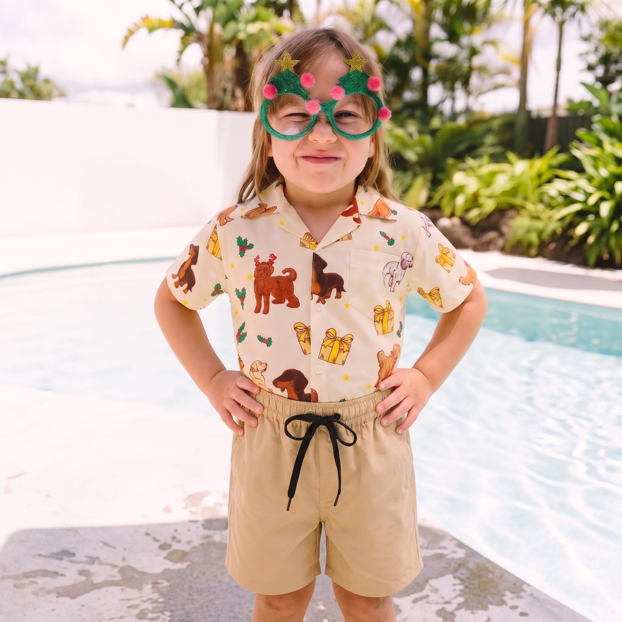 A young girl stands in front of a swimming pool wearing a short-sleeve button-up shirt with a beige background and a colorful print of cartoon dogs, cacti, and hot air balloons, paired with beige shorts and novelty green glasses decorated with pink stars.