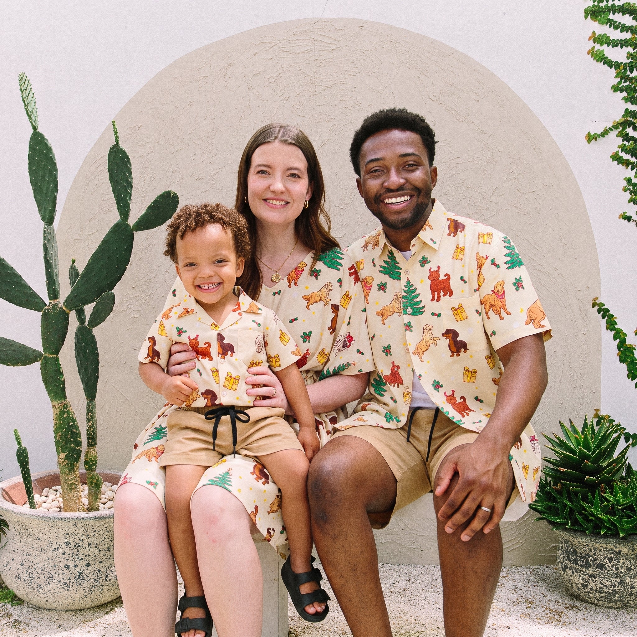 A family of three wearing matching beige short-sleeve button-up shirts and shorts sets with a colorful dog-themed print, posing together in front of a white arched wall with potted cacti.